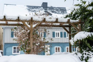 Eine schneebedeckte Holzpergola steht vor einem blau-grauen Haus mit weißem Schnee auf dem Dach und den Fensterläden sowie schneebedeckten Bäumen und Büschen im Hof.