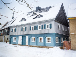 Ein traditionelles zweistöckiges Haus mit blau-grauer Außenverkleidung, weißen Zierleisten und schneebedecktem Dach steht an einer verschneiten Straße – eine einladende Ferienwohnung in der Oberlausitz für alle, die einen gemütlichen Aufenthalt im Herzen Sachsens suchen.
