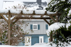 Eine schneebedeckte Holzpergola umrahmt den Blick auf ein blau-graues Haus mit weiß eingefassten Fenstern, blauen Fensterläden und einer schneebedeckten Fichte und Büschen im Vordergrund.