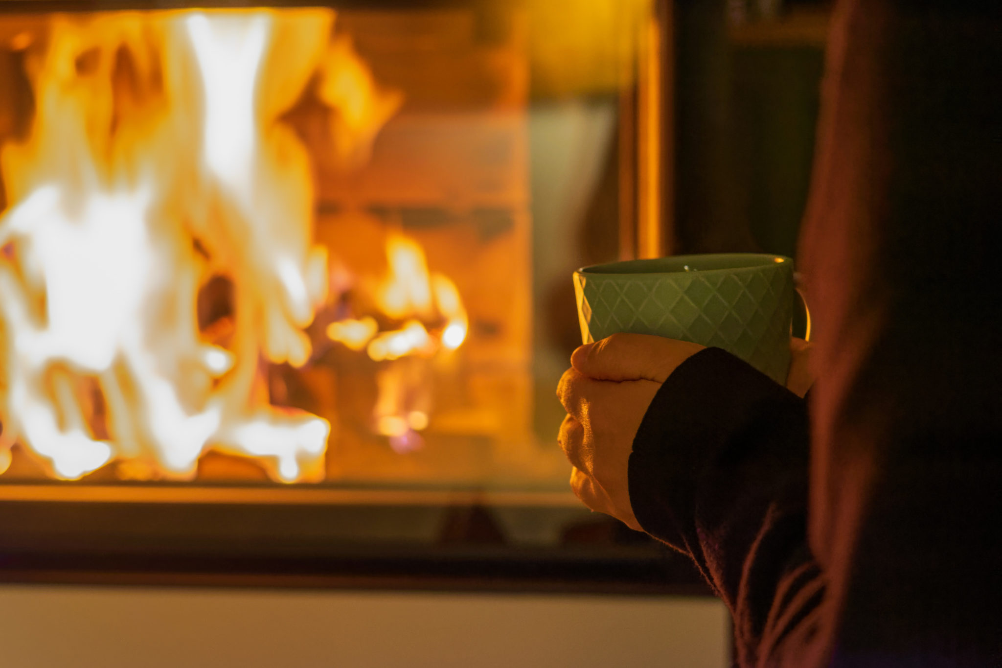 Eine Person mit einem grünen Becher steht neben einem Kamin und wärmt sich an den Flammen. Die Szene ist gemütlich und sanft beleuchtet, was die Wärme und Behaglichkeit des Feuers unterstreicht.
