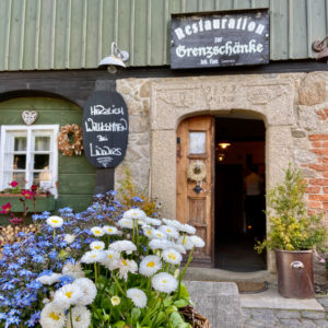 Lindners Grenzschenke ist ein rustikaler Restauranteingang mit Steinmauern und Holztüren, der mit Blumenkränzen und Topfblumen besticht. Ein Kreideschild begrüßt die Besucher - ein echter Gastrotipp. Im Vordergrund steht ein Korb mit weißen und blauen Blumen.