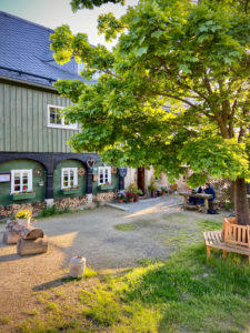 Ein charmantes Landhaus in der Oberlausitz mit grünen Holzwänden und weißen Fenstern, geschmückt mit Topfpflanzen und Kränzen. Ein belaubter Baum beschattet eine Holzbank und einen Picknicktisch - perfekt für den Genuss in Lindners Grenzschänke.