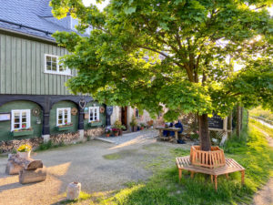 Ein großer Baum mit einer Holzbank um den Stamm herum steht vor einem rustikalen Umgebindehaus mit weiß umrandeten Fenstern; an einem Picknicktisch in der Nähe des Eingangs sitzen Menschen, umgeben von Pflanzen und Sonnenlicht - ideal für genussvollen Urlaub in der Oberlausitz. Hervorragendes Restaurant "Lindners Grenzschänke" in Friedersdorf.