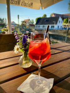 Ein Glas roter Cocktail mit Eis und einem Strohhalm steht auf einem Holztisch neben Wildblumen, draußen an einem sonnigen Tag - Genuss pur in der Oberlausitz, mit Häusern und üppigem Grün im Hintergrund.