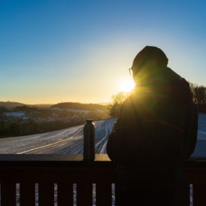 Eine Person in Wintermantel und Mütze steht bei Sonnenuntergang auf einer Aussichtsplattform in Neusalza-Spremberg, mit Blick auf ein verschneites Feld und entfernte Bäume, mit einer Thermoskanne auf dem Geländer und dem Sonnenlicht im Rücken.