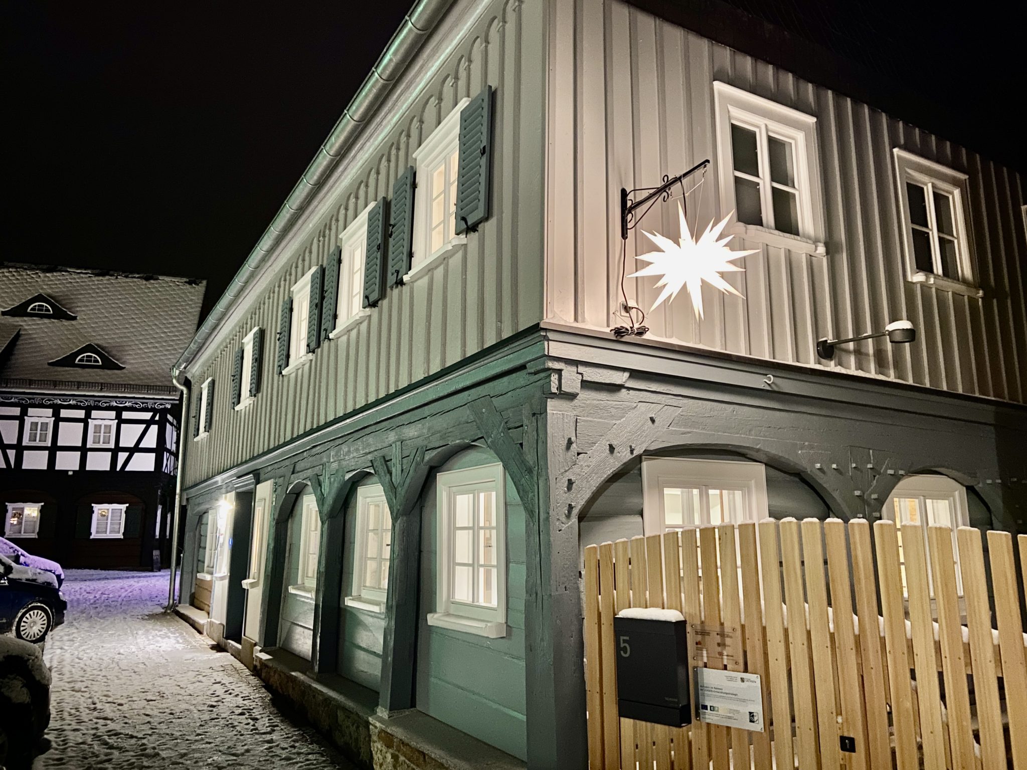 Ein zweistöckiges Fachwerkhaus mit blauen Fensterläden und weiß gerahmten Fenstern ist nachts beleuchtet und vermittelt den Charme eines entspannten Winterurlaubs. Eine leuchtender Herrnhuter Stern hängt an der Fassade, Schnee bedeckt die Dächer, und im Vordergrund stehen ein Holzzaun und ein Briefkasten.