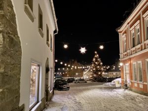 Eine verschneite Straße in Neusalza-Spremberg ist nachts mit Lichterketten und Herrnhuter Sternen geschmückt. Ein großer Weihnachtsbaum mit Lichtern steht neben geparkten Autos, und die festlichen Fenster des Obermarkts erhellen die Szene.