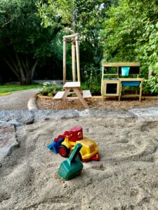 Spielzeug-Baufahrzeuge und eine grüne Schaufel stehen in einem sandigen Spielbereich, mit einem hölzernen Picknicktisch, einer Spielküche und üppigen grünen Bäumen im Hintergrund an einem hellen Tag.