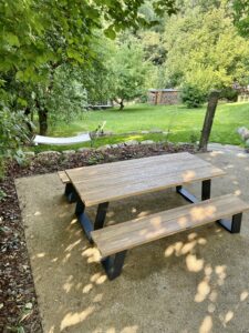 Ein hölzerner Picknicktisch mit Bänken steht auf einer Kiesfläche unter grünen Bäumen, mit Blick auf eine grüne Wiese mit einer Hängematte und gestapeltem Brennholz - perfekt für einen entspannten Urlaub in Sachsen im charmanten Neusalza-Spremberg.
