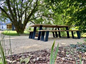 Ein hölzerner Picknicktisch mit schwarzen Metallbeinen steht auf einem Kiesweg unter grünen Bäumen in einem sonnenbeschienenen Park, mit einem Ferienhaus und Garten im Hintergrund - perfekt für Urlaub in der Oberlausitz.