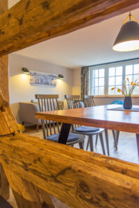 Der Essbereich in der Ferienwohnung Hermann mit einem Holztisch und grauen Stühlen, der durch rustikale Holzbalken zu sehen ist; ein Sofa, Wandkunst, ein großes Fenster und eine Hängelampe sind in dem gemütlichen, gut beleuchteten Raum zu sehen.