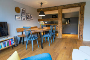 Der gemütliche Essbereich in der Ferienwohnung Hermann mit einem Holztisch, blauen Stühlen, einer Bank und rustikalen Balken - perfekt für eine Ferienwohnung für große Familien. Der Raum ist mit einer modernen Küche mit dunklen Schränken verbunden. Ein Fernseher, ein Bücherregal und Wandkunst verleihen dem Raum eine warme Atmosphäre.