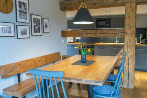 Ein gemütlicher Essbereich in einer Ferienwohnung für Familienurlaub mit einem Holztisch, zwei blauen Stühlen und einer Holzbank in der Nähe einer Küche mit grauen Schränken. Eine schwarze Hängelampe hängt über dem Tisch, und in der Mitte stehen Narzissen in einer Vase.
