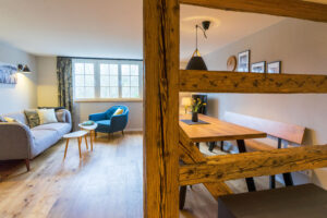 Ein gemütlicher Wohn- und Essbereich, der durch Holzbalken unterteilt ist, mit einem grauen Sofa, einem blauen Sessel, hölzernen Kaffee- und Esstischen, Stühlen, Sitzbänken und großen Fenstern, die natürliches Licht hereinlassen.