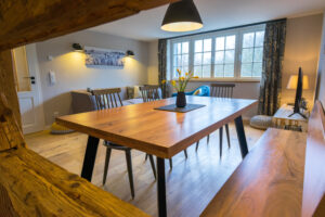 Das gemütliche Esszimmer im Ferienhaus Marktquartier 5 besteht aus einem Holztisch mit Bänken, modernen Stühlen, einer schwarzen Pendelleuchte, großen Fenstern mit Vorhängen und einer Vase mit gelben Blumen. Das natürliche Licht füllt den Raum und schafft eine warme Atmosphäre.