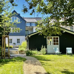 Ein schwarzes Gartenhaus mit weißer Tür und zwei blauen Stühlen steht in einem üppigen Garten. Im Hintergrund ist ein großes weiß-blaues Haus unter einem strahlend blauen Himmel zu sehen. Ein gewundener Pfad führt durch den grasbewachsenen Hof.