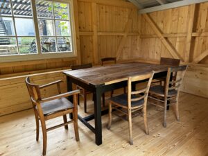 Ein Holztisch mit sechs nicht zusammenpassenden Stühlen steht in einem sonnenbeschienenen Gartenhaus mit Holzwänden und -boden. Ein großes Fenster füllt den Raum mit natürlichem Licht und gibt den Blick frei auf die Gartenmöbel und die Terrasse dahinter. Perfekt für Workation oder Urlaub in der Oberlausitz.