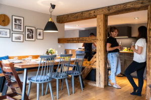 Zwei Frauen unterhalten sich bei einem Getränk in einem gemütlichen, modernen Essbereich einer Ferienwohnung in der Oberlausitz mit Holzbalken und einem gedeckten Esstisch, während ein Mann im Hintergrund das Essen zubereitet - perfekt für Urlaub in der Oberlausitz.