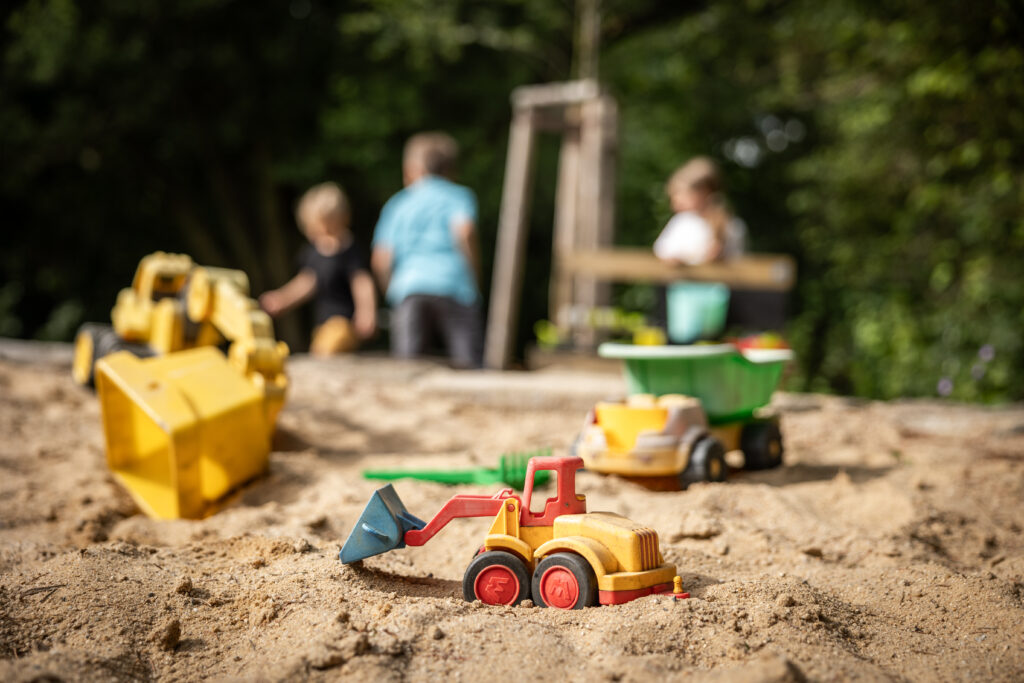 Spielzeug-Baufahrzeuge in einem Sandkasten mit drei spielenden Kindern im unscharfen Hintergrund. Die Szene ist im Freien mit grünen Bäumen und Sonnenlicht. Der malerische Ort befindet sich direkt hinter dem Marktquartier 5 mit drei Ferienwohnungen in Sachsen.