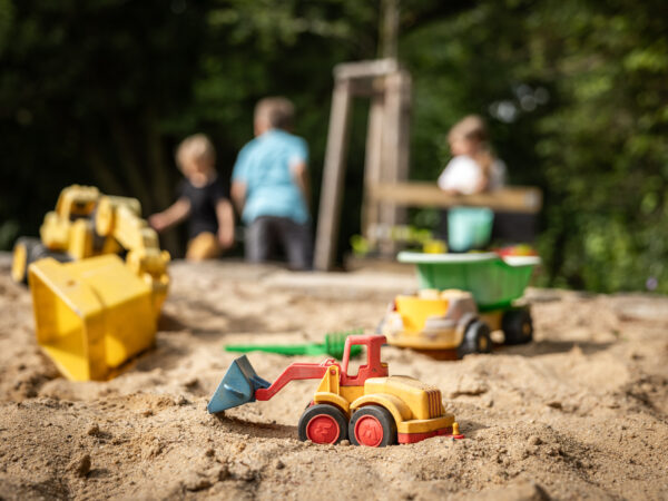 Spielzeug-Baufahrzeuge in einem Sandkasten mit drei spielenden Kindern im unscharfen Hintergrund. Die Szene ist im Freien mit grünen Bäumen und Sonnenlicht. Der malerische Ort befindet sich direkt hinter dem Marktquartier 5 mit drei Ferienwohnungen in Sachsen.
