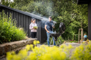 Eine Frau und ein Mann bereiten in einem üppigen Garten an einem Holzkohlegrill Essen zu. Der Mann grillt, während die Frau eine Schüssel Salat hält. Rauch steigt vom Grill auf, Pflanzen und Blumen säumen die gemauerte Terrasse. Der malerische Ort befindet sich direkt hinter dem Marktquartier 5, einem Ferienhaus mit Garten in Neusalza-Spremberg.