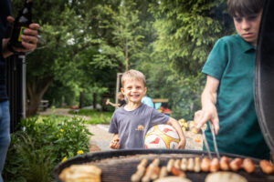 Ein lächelnder Junge mit einem Fußball steht neben einem Grill, auf dem eine andere Person Würstchen grillt. Im Hintergrund sind grüne Bäume und ein Picknicktisch zu sehen.