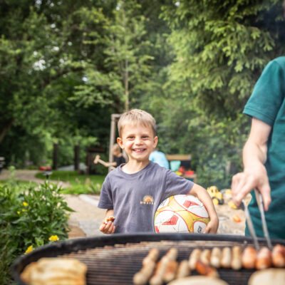 Ein lächelnder Junge mit einem Fußball steht neben einem Grill, auf dem eine andere Person Würstchen grillt. Im Hintergrund sind grüne Bäume und ein Picknicktisch zu sehen.
