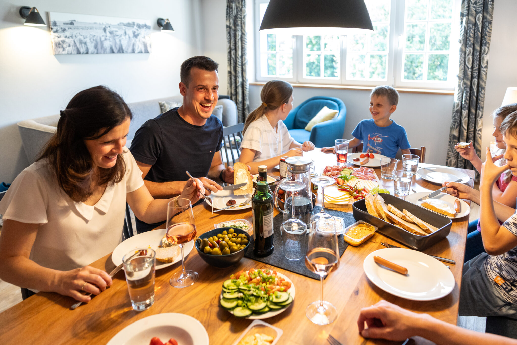 Eine sechsköpfige Familie sitzt um einen hölzernen Esstisch in ihrer Ferienwohnung in der Oberlausitz und teilt lächelnd und lachend eine Mahlzeit mit Brot, Oliven, Käse und frischem Gemüse. Die gemütliche Umgebung fängt die Freude am Urlaub in der Oberlausitz ein.
