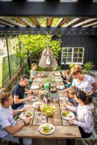 Eine Familie versammelt sich um einen Holztisch im Freien ihres Ferienhauses mit Garten und isst unter einer Pergola mit Hängelampen. Teller mit Essen, Getränken und Salat stehen bereit, umgeben von Grün und einem schwarzen Gartenhaus im Hintergrund.