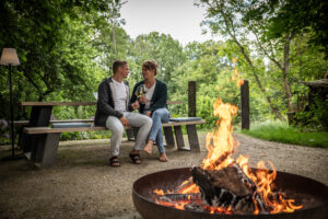 Ein Paar sitzt auf einer Holzbank im üppigen Garten ihres Ferienhauses mit Garten und lächelt sich mit Weingläsern in der Hand an, während im Vordergrund eine warme Feuerstelle brennt und sie von Bäumen umgeben sind.