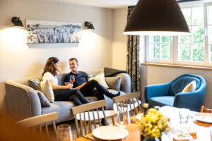 Ein Paar sitzt auf einem grauen Sofa, lächelt und hält Weingläser in einer hellen, gemütlichen Ferienwohnung für große Familien mit großen Fenstern, einem blauen Sessel und einem gedeckten Esstisch im Vordergrund.
