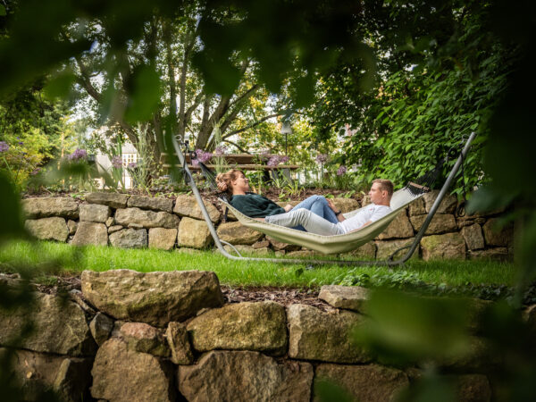 Ein Paar entspannt und unterhält sich, während es zusammen in einer Hängematte in einem üppigen, grünen Garten liegt, umgeben von Bäumen und Steinmauern. Der malerische Ort befindet sich direkt hinter dem Marktquartier 5, einem Ferienhaus mit Garten in Neusalza-Spremberg.