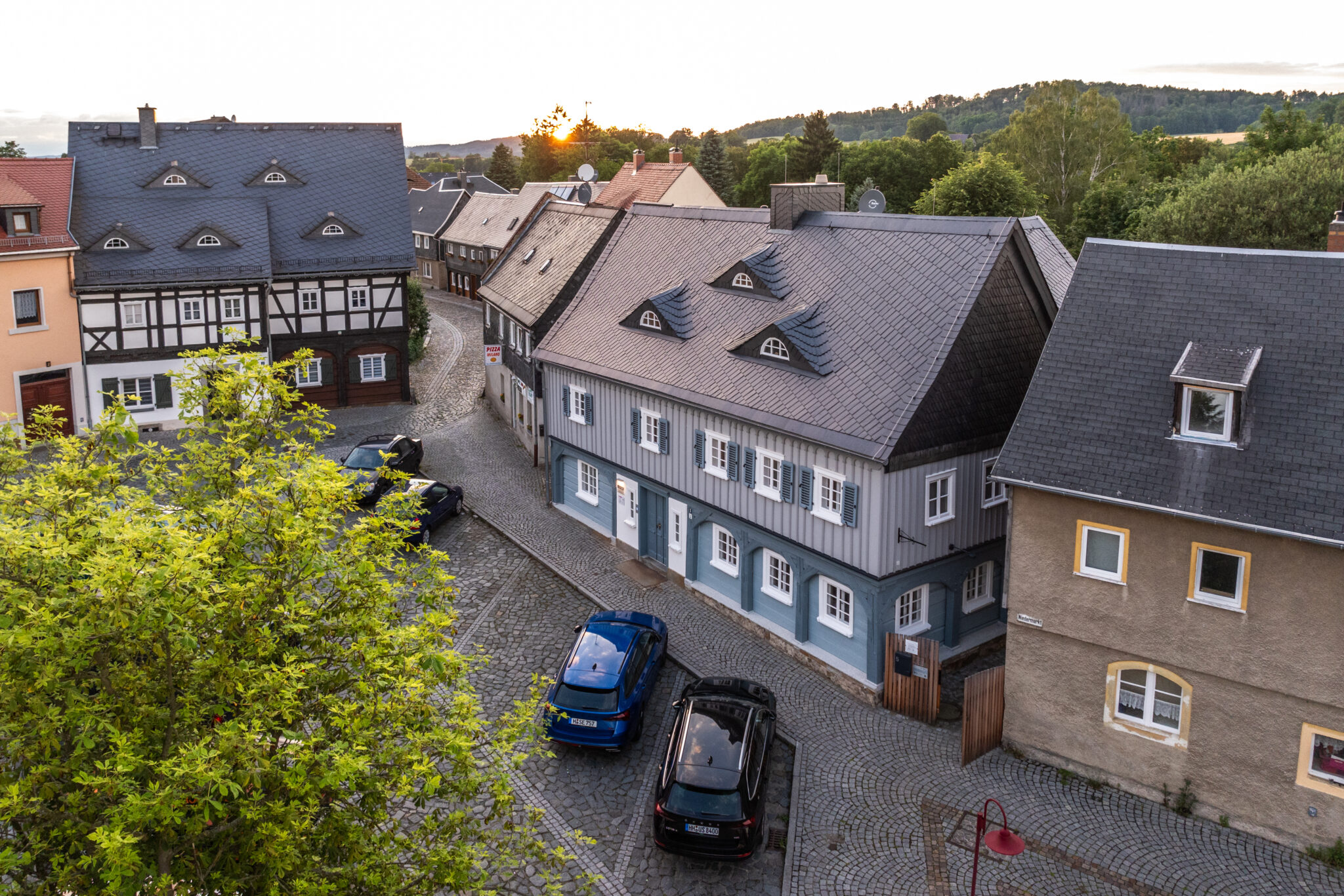 Eine ruhige Kopfsteinpflasterstraße, gesäumt von traditionellen europäischen Häusern, darunter ein charmantes Umgebindehaus – perfekt für „Urlaub im Umgebindehaus“ – mit am Straßenrand geparkten Autos und grünen Bäumen im Vordergrund bei Sonnenuntergang.