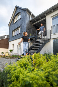 Eine Frau und ein Mann gehen eine Metalltreppe im Freien hinunter, die zu einem modernen Haus mit dunkelblauer Außenverkleidung und weißen Wänden führt, umgeben von Grün und gelben Blumen.