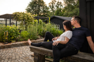 Ein Paar sitzt eng beieinander auf einer Steinbank in einem Garten, umgeben von üppigem Grün und Holzgebäuden, und genießt einen ruhigen Moment im Freien am Abend.