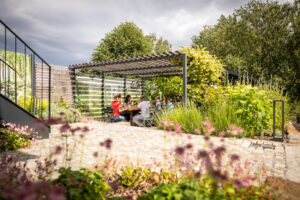 Menschen sitzen und unterhalten sich unter einer Pergola in einem üppigen, sonnigen Garten, umgeben von Blumen, Sträuchern und Bäumen, über dem ein bewölkter Himmel hängt. Der Ort befindet sich direkt hinter dem Marktquartier 5, einem Ferienhaus mit Garten in Neusalza-Spremberg.