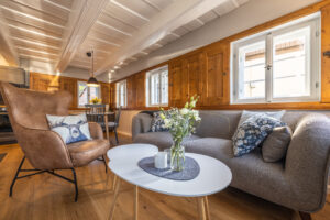 Helles Wohnzimmer mit einem modernen grauen Sofa, gemusterten Kissen und einem weißen Couchtisch mit Blumen. Große Fenster mit Holzläden füllen diese Ferienwohnung in der Oberlausitz mit Licht; im Hintergrund ist der Essbereich sichtbar.