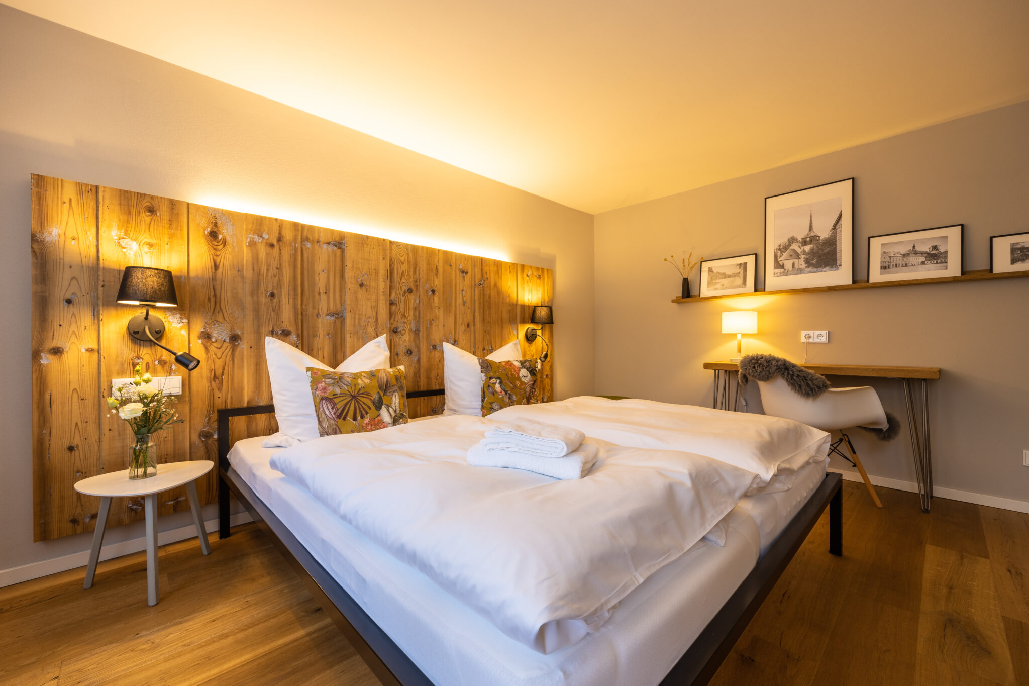 Ein gemütliches, modernes Schlafzimmer mit Holzboden, einem Doppelbett mit weißer Bettwäsche und zwei dekorativen Kissen. Die Ferienwohnung mit Sauna verfügt über ein rustikales Holzkopfteil mit montierten Lampen sowie einen kleinen Schreibtisch, einen Stuhl und gerahmte Kunst an der gegenüberliegenden Wand. Großes Schlafzimmer im Apartment Alwine im Ferienhaus Marktquartier 5.