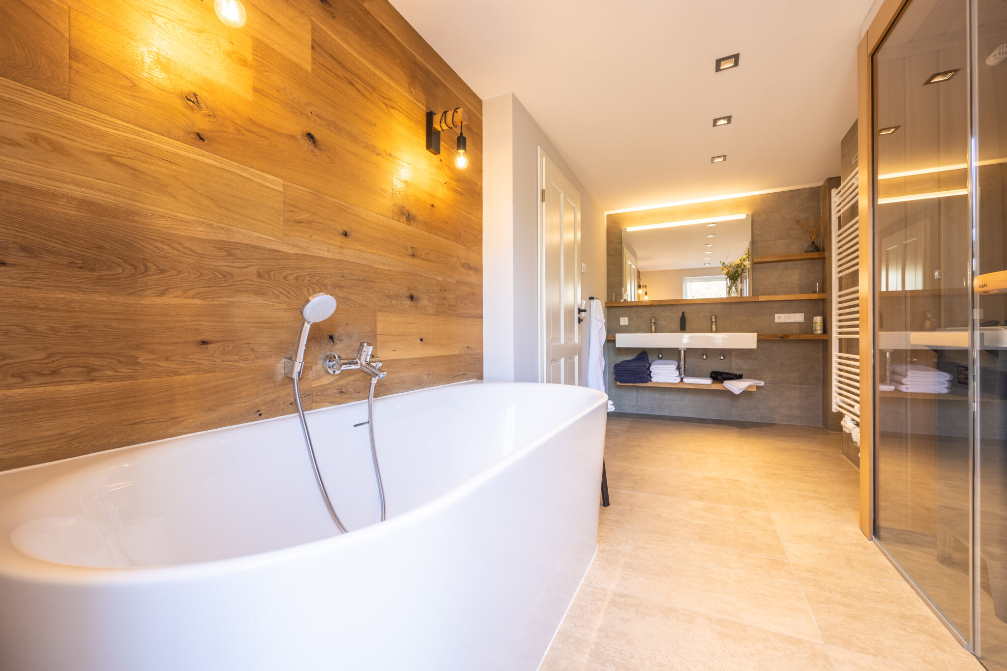 Modernes Badezimmer in einer Ferienwohnung in der Oberlausitz, ausgestattet mit einer großen weißen Badewanne, einer holzgetäfelten Wand, einer Sauna, einem Doppelwaschbecken mit beleuchtetem Spiegel, Regalen mit Handtüchern und neutralem Fliesenboden für einen entspannenden Rückzugsort. Bad im Ferienhaus Marktquartier 5 in Neusalza-Spremberg