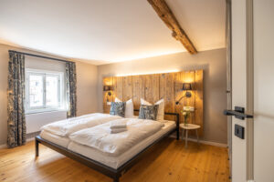 Ein gemütliches Schlafzimmer in der Ferienwohnung Hermann im Marktquartier 5 für große Familien mit einem Doppelbett, weißen Bettwaren, gemusterten Kissen, rustikalem Holzkopfteil, zwei Wandlampen, kleinem runden Tisch mit Blumen, Holzboden und Fenster mit Vorhängen.