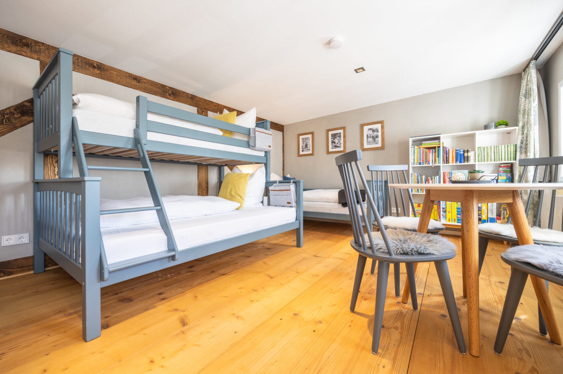 Ein helles Schlafzimmer in einer komfortablen Ferienwohnung für große Familien mit hellen Holzfußböden, blauen Etagenbetten, Einzelbett, rundem Tisch und Bücherregalen. Große Fenster lassen viel Tageslicht herein. Spiele und Bücher versprechen Spaß und Abwechslung für den Familienurlaub in der Oberlausitz.