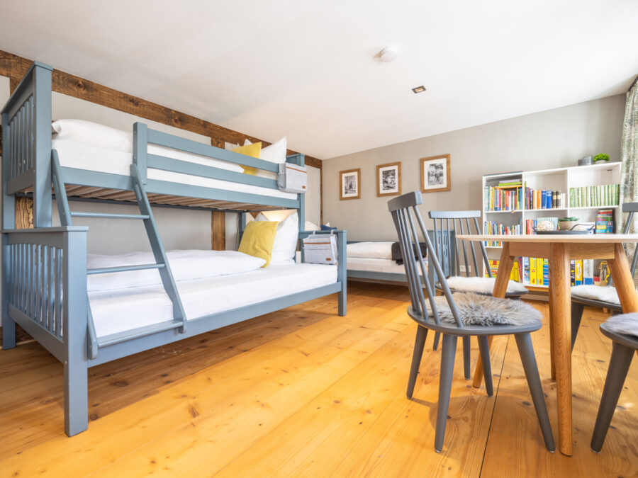 Ein helles Schlafzimmer in einer komfortablen Ferienwohnung für große Familien mit hellen Holzfußböden, blauen Etagenbetten, Einzelbett, rundem Tisch und Bücherregalen. Große Fenster lassen viel Tageslicht herein. Spiele und Bücher versprechen Spaß und Abwechslung für den Familienurlaub in der Oberlausitz.