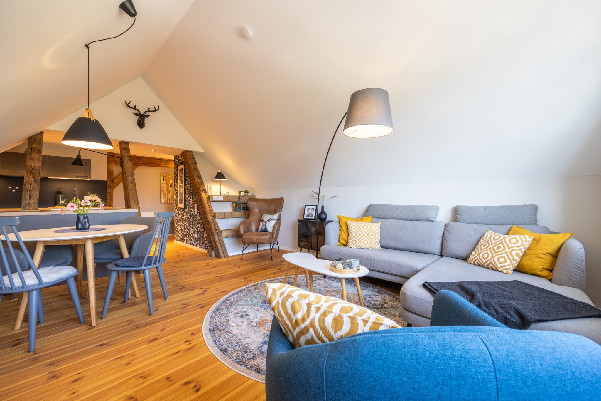 Ein gemütliches, modernes Dachgeschoss-Wohnzimmer mit Holzfußboden, einem grauen Sektionssofa mit gelb gemusterten Kissen, einem runden Teppich, einem blauen Sessel, einer Stehlampe, einem Essbereich und freiliegenden Holzbalken. Willkommen in unserer Ferienwohnung Carl für zwei Personen!
