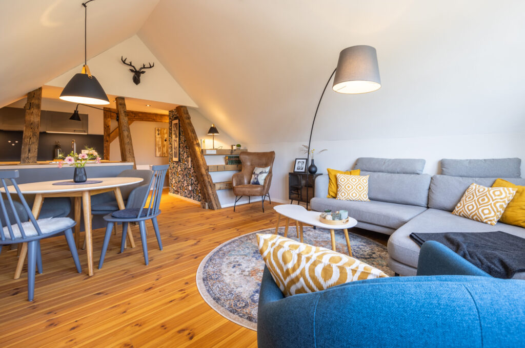 Ein gemütliches, modernes Dachgeschoss-Wohnzimmer mit Holzfußboden, einem grauen Sektionssofa mit gelb gemusterten Kissen, einem runden Teppich, einem blauen Sessel, einer Stehlampe, einem Essbereich und freiliegenden Holzbalken. Willkommen in unserer Ferienwohnung Carl für zwei Personen im Herzen der Oberlausitz!