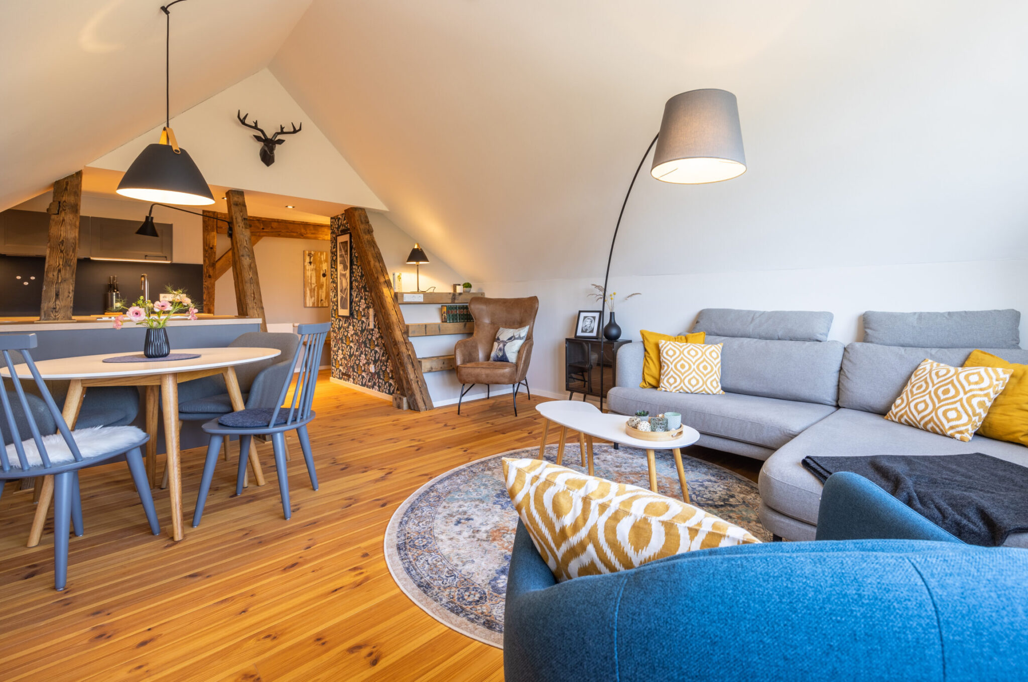 Ein gemütliches, modernes Dachgeschoss-Wohnzimmer mit Holzfußboden, einem grauen Sektionssofa mit gelb gemusterten Kissen, einem runden Teppich, einem blauen Sessel, einer Stehlampe, einem Essbereich und freiliegenden Holzbalken. Willkommen in unserer Ferienwohnung Carl für zwei Personen im Herzen der Oberlausitz!