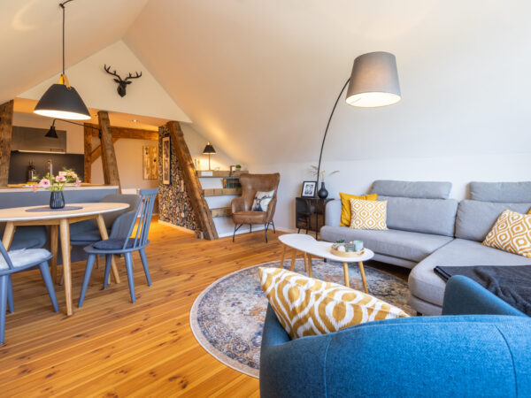 Ein gemütliches, modernes Dachgeschoss-Wohnzimmer mit Holzfußboden, einem grauen Sektionssofa mit gelb gemusterten Kissen, einem runden Teppich, einem blauen Sessel, einer Stehlampe, einem Essbereich und freiliegenden Holzbalken. Willkommen in unserer Ferienwohnung Carl für zwei Personen im Herzen der Oberlausitz!