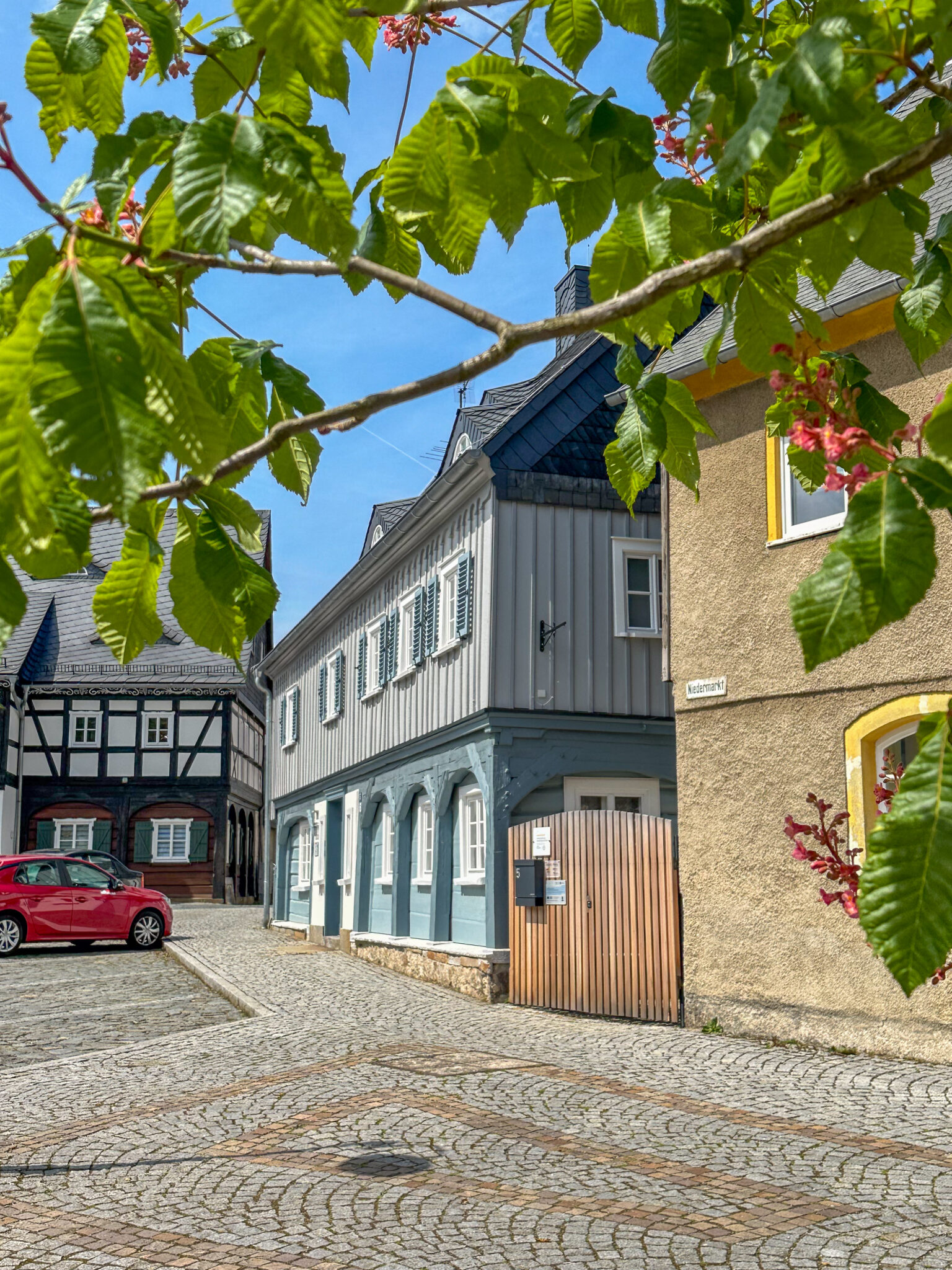Eine Kopfsteinpflasterstraße mit traditionellen Fachwerkhäusern und bunten Häusern, links parkt ein rotes Auto und im Vordergrund grüne Blätter mit rosa Blüten unter einem blauen Himmel.