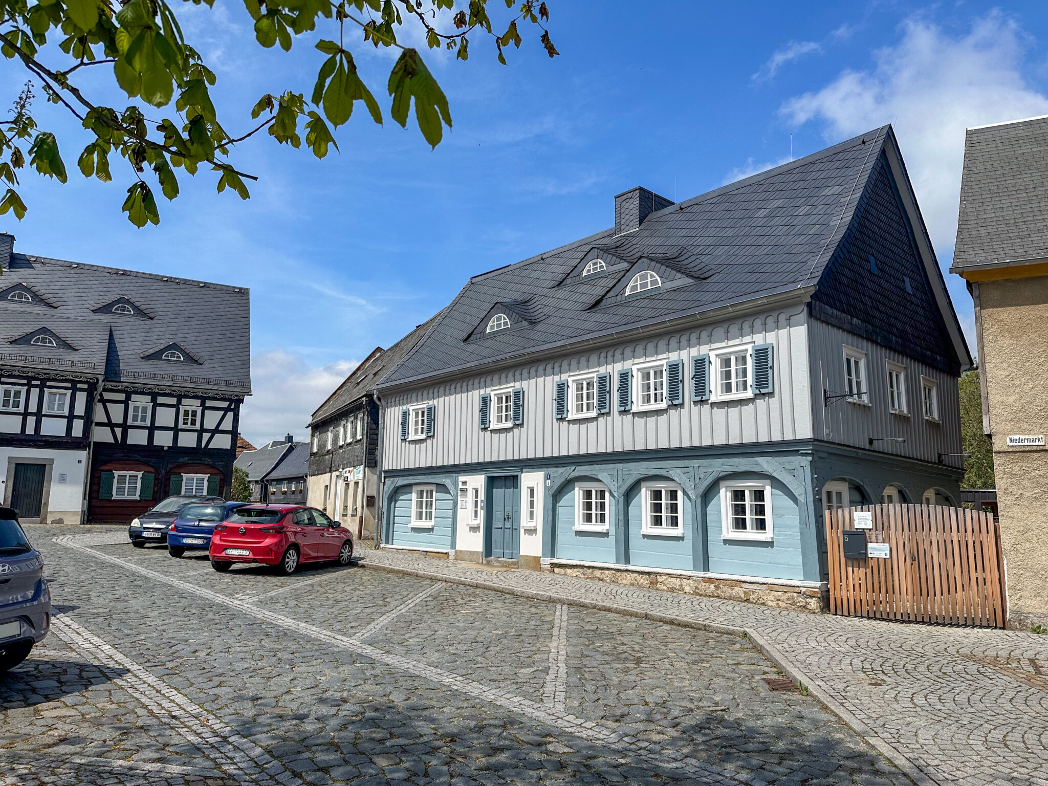 Eine Kopfsteinpflasterstraße mit bunten Fachwerkhäusern, darunter ein großes blaugraues mit weißen Fensterläden: Das Marktquartier 5 - Ort für Urlaub im Denkmal. Mehrere geparkte Autos säumen die Straße unter einem sonnigen, teilweise bewölkten Himmel.
