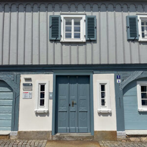 Ein blau-weißes Fachwerkhaus mit einer zentralen Doppeltür, zwei kleinen Fenstern auf beiden Seiten und einem größeren Fenster mit Fensterläden darüber. Der Eingang ist von Kopfsteinpflaster eingerahmt. Ein typisches oberlausitzer Umgebindehaus.