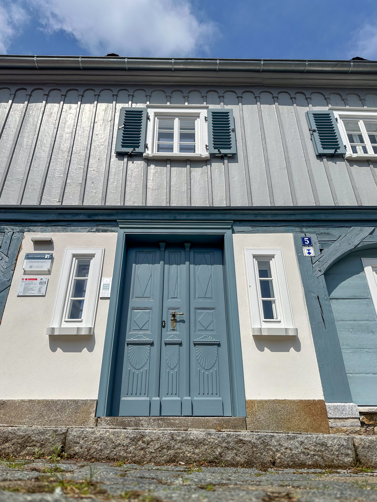 Eine Untersicht auf ein traditionelles Umgebindehaus mit einer blauen Doppeltür, weißen Wänden, kleinen Fenstern mit Fensterläden und einer Steinstufe am Eingang unter einem teilweise bewölkten Himmel.